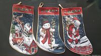 Three Christmas stockings arranged side by side on a dark surface, showing full front embroidery detail of Santa with presents, snowman, and Santa sleigh with reindeer.