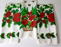 Three Christmas-themed tea towels folded to show poinsettia and holly leaf designs