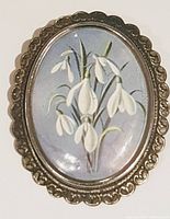 Front view of oval silver-toned brooch with painted snowdrop flower design on blue background and scalloped border.