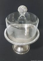 Two piece dessert stand set shown assembled. Silvertone round pedestal base with crystal rhinestone detail around rim. Clear glass dome with crystal rhinestone knob on top.