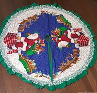 Full view of circular Christmas tree skirt laid flat, showing entire Santa Claus design and green ruffle with white lace trim