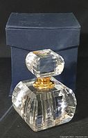 Front view of the lead crystal perfume bottle with its square faceted stopper and gold-tone neck collar, displayed in front of the original navy blue box.