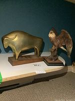 Photo of both brass bull figure and resin eagle figure side by side on a white surface against a blue background.