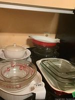 Overview of various glass and ceramic bakeware including Pyrex cherry-pattern bowls, ceramic casserole dishes, and clear glass casseroles and baking dishes stacked on a stove.
