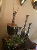 Photo showing large brass style lamp with harp and two tall metal vases behind it on a wooden table