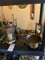 Photo showing assorted brass and metal decorative items including candle lantern, trivet, flower pot, napkin rings, and metal bottles.