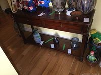 Full view of the wooden console table with two drawers, lower shelf, and tabletop showing wear and discoloration. Various decorative items on and around table are not included.