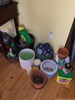 Overview of assorted gardening items including watering globes, various ceramic and plastic planters, and plant food containers on floor near wall and furniture