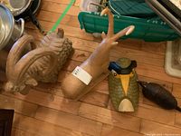 Photo showing two mountain goat figurines side by side, one with curled horns and wool texture, the other smoother with a damaged horn. Also visible are a decorative owl mask and a smaller armadillo figurine alongside them on a wooden floor.