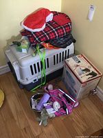 Photo showing the large plastic dog kennel with red and black plaid and gray dog coats on top, box with pet drinking fountain, metal bowl and dog toys on floor.