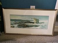 Framed wide horizontal print of a polar bear on snowy ice looking out over a rocky shore with water beyond. Gold-tone frame and white matting.