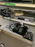 Shelving with PS2 console, controllers on charging dock, 3 PS2 games stacked, and Sony SLV-N81 VCR with some cables on top.