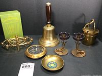 All seven brass and glass decorative items grouped on a black backdrop: two flower candle holders with amber glass cups, large town crier bell with wooden handle, brass bucket with lid and handle, small brass dish with blue enamel center, glass coaster with brass rim etched 1955, and Colonial Candle Co. oval candle holder with original green box.
