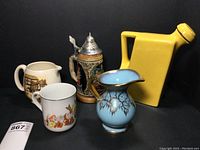 All five ceramic vessels shown together against black background: yellow Oxford Ware pitcher, Lancaster & Sandland English pitcher, blue and gold German pitcher, German beer stein with metal lid, and Snow White mug.
