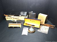 Full lot arranged showing Buck knife with leather sheath and original box, silver tone flask, Waterman fountain pen in box, Koh-i-noor Radiograph pen in box, glass magnifying paperweight, uncirculated stamps, leather riding crop, enamel decorative pipe