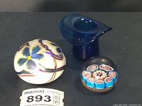 Three art glass paperweights: two Orient & Flume floral tiger lily paperweights and one blue glass hat by Piedmont Art Glass, with small millefiori paperweight in front.