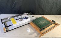 Four paper cutters and trimmers displayed on table including wooden guillotine cutter, boxed Promaster DigiTrim Rotary Trimmer, and two stainless steel Rodafilm trimmers.