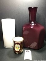 Three glass vases: white cylindrical vase, purple square carafe style vase, and short hand painted vase
