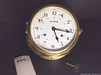 Front view of the brass Royal Mariner ships clock with winding key beside it, showing clear dial markings and hands.