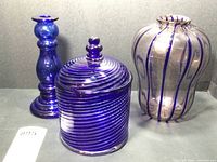 Three blue glass decor items including candle holder, swirl jar with lid, and striped vase