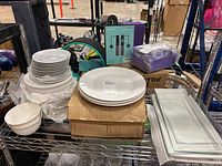 Overview photo showing stacks of plates, bowls, and rectangular platters on a metal rack with cardboard boxes and other items in the background.