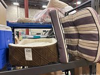 Brown plush dog bed with quilted pattern and striped patio lounge chair cushion on metal shelf.