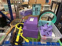 Photo showing two purple yoga blocks stacked, digital jump rope in turquoise and black box, two vintage wooden tennis racquets, a green exercise wheel frame, and a plastic bin containing fitness accessories