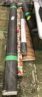 Five rolls of flooring and wall covering leaning against a table and resting on a rough concrete floor, showing the various textures and patterns including grey stone look, wood grain, floral, and grass cloth texture.