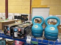 Pair of blue adjustable kettlebell weights with multiple weight plates and two boxed Shake Weights on shelf