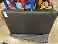Folded black massage table standing upright with attached handle and black protective cover partially unzipped at bottom exposing table edge.