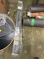 Metal shelving parts wrapped in plastic and secured with tape, laid on floor with rolled rugs nearby.