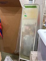 Freestanding full length mirror standing upright next to cardboard and plastic wrap in warehouse setting.