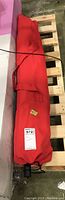 Red fabric carry case for umbrella, rolled up and secured with red straps, stored on a wooden pallet.