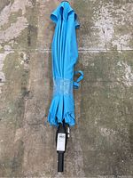 Top view of folded blue patio umbrella canopy with strap securing it, showing the black umbrella frame's pole base without the pole itself.