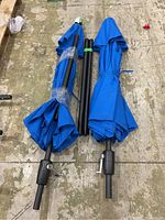 Two folded blue patio umbrella canopies, each attached to black poles, laid side by side on a textured floor. One pole has a crank handle for operation and the umbrellas have black pole segments wrapped together.