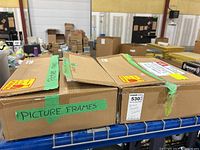 Two closed cardboard boxes labeled 'Picture Frames' on a metal shelf in a warehouse setting.