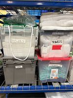 Four stacked plastic bins labeled 'Towels' holding white and blue towels, some towels folded on top, stored on metal shelving.
