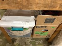 Photo of two storage bins (one plastic with lid, one cardboard) containing towels and bath mats under a wooden table.