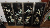 Four black lacquer wooden panels with intricate mother of pearl inlays of birds and flowers arranged side by side on a floor.