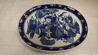 Full top view of the oval blue and white painted ceramic plate showing the scene of traditional figures and border pattern.