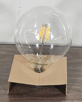 Clear glass G125 shaped light bulb showing yellow LED filament inside, seated on brown cardboard holder.