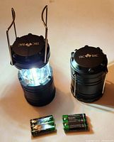 Photo showing 2 IBC branded compact lanterns, one lit and extended, the other collapsed, with included batteries