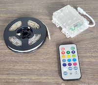 RGB LED strip light on black spool, transparent waterproof battery box with attached cable, and white remote control with color buttons laid on wooden surface.