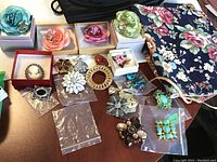 Overview photo of approximately 19 brooches arranged on table with two floral fabric pouch bags beside them