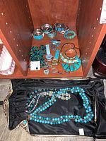 View showing assorted costume jewelry items including turquoise and blue bracelets, watches, earrings, and necklaces arranged in shelving cubby and a black bag.