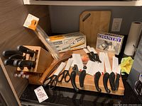 Full view of wooden knife block with several knives, multiple black-handled cutters/scissors laid out, a wooden cutting board in the background, and boxed items on counter.