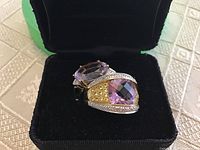 Two rings in black presentation box, angled view showing checkerboard cushion amethyst ring in front and oval amethyst ring behind.