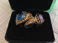 Two gold rings displayed side by side in black velvet case showing blue topaz and red garnet stones