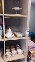 Full view of tea set and serving items displayed on shelving, showing cups, saucers, cream, sugar, plates, and tiered serving stand.