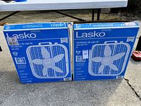 Two boxed Lasko air circulating box fans, shown outdoors on pavement next to a table, both boxes sealed with handle cutout visible.